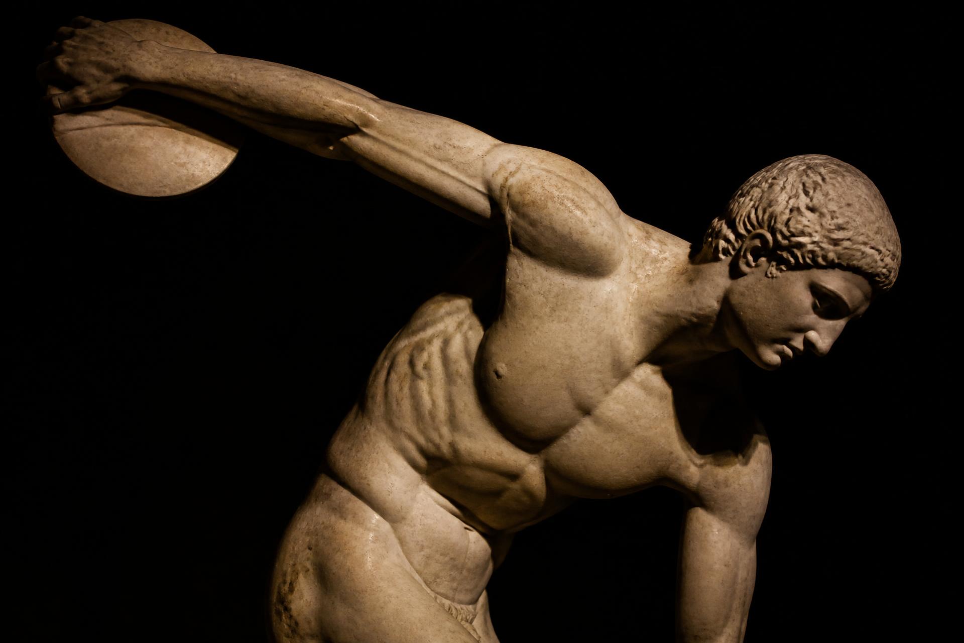 Classical marble sculpture of a discus thrower, arm extended, lit against a dark ground
